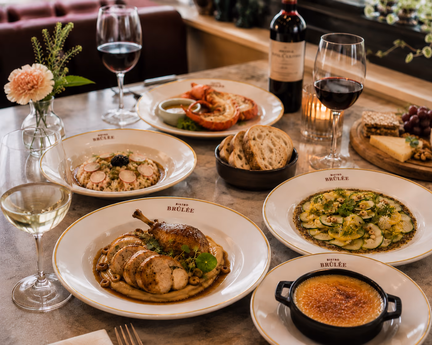 Table with plated gourmet dishes, wine glasses, bread, cheese, grapes, and a flower vase at Bistro Brûlée.