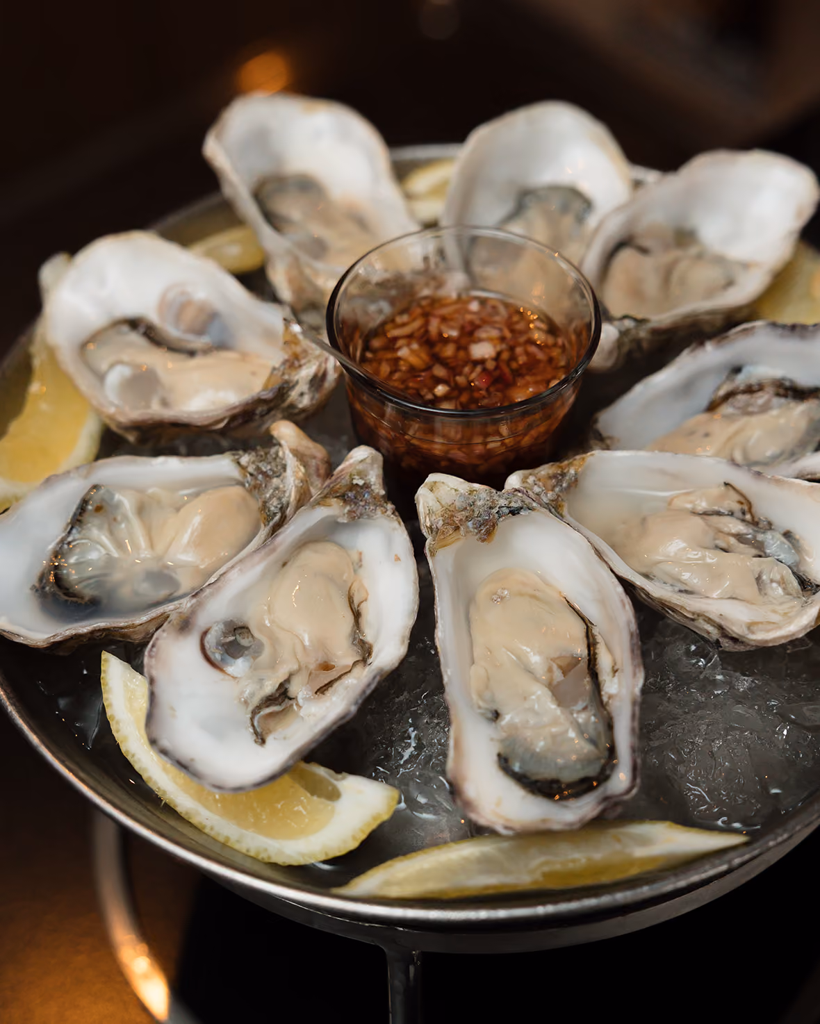 Tray of fresh oysters on ice with lemon wedges and a cup of dipping sauce in the center.