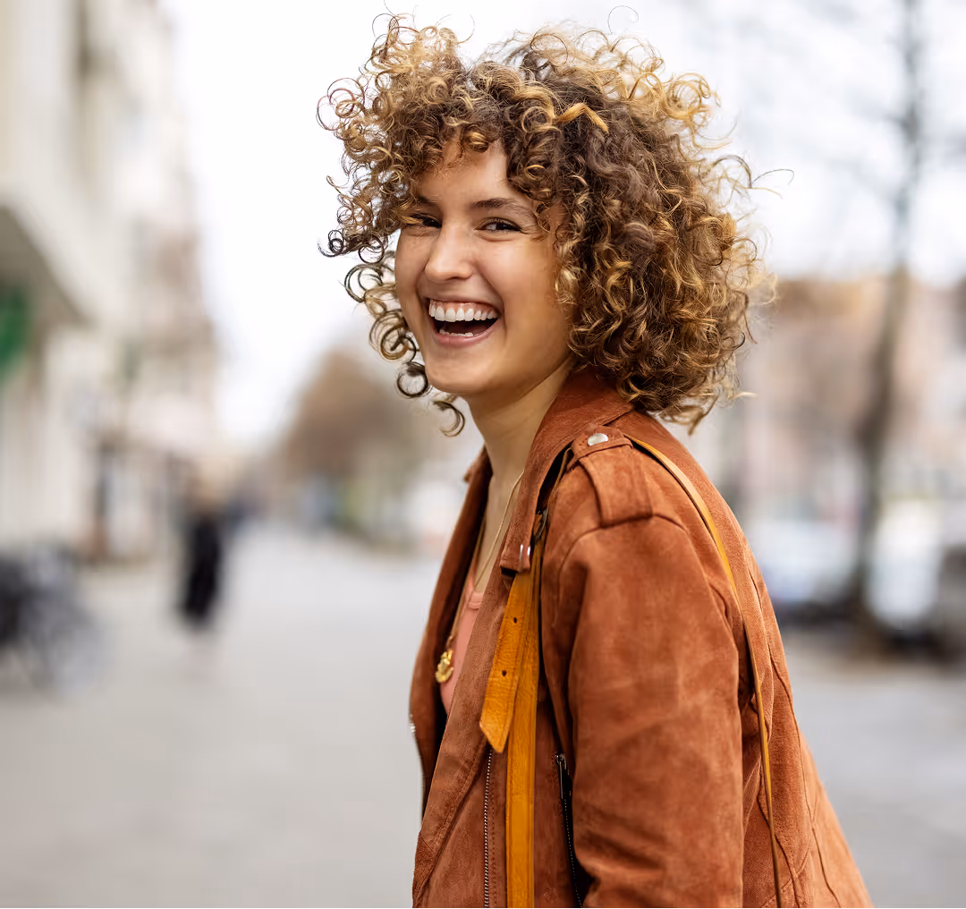 Smiling woman with curly hair wearing a brown jacket standing on a blurred city street.