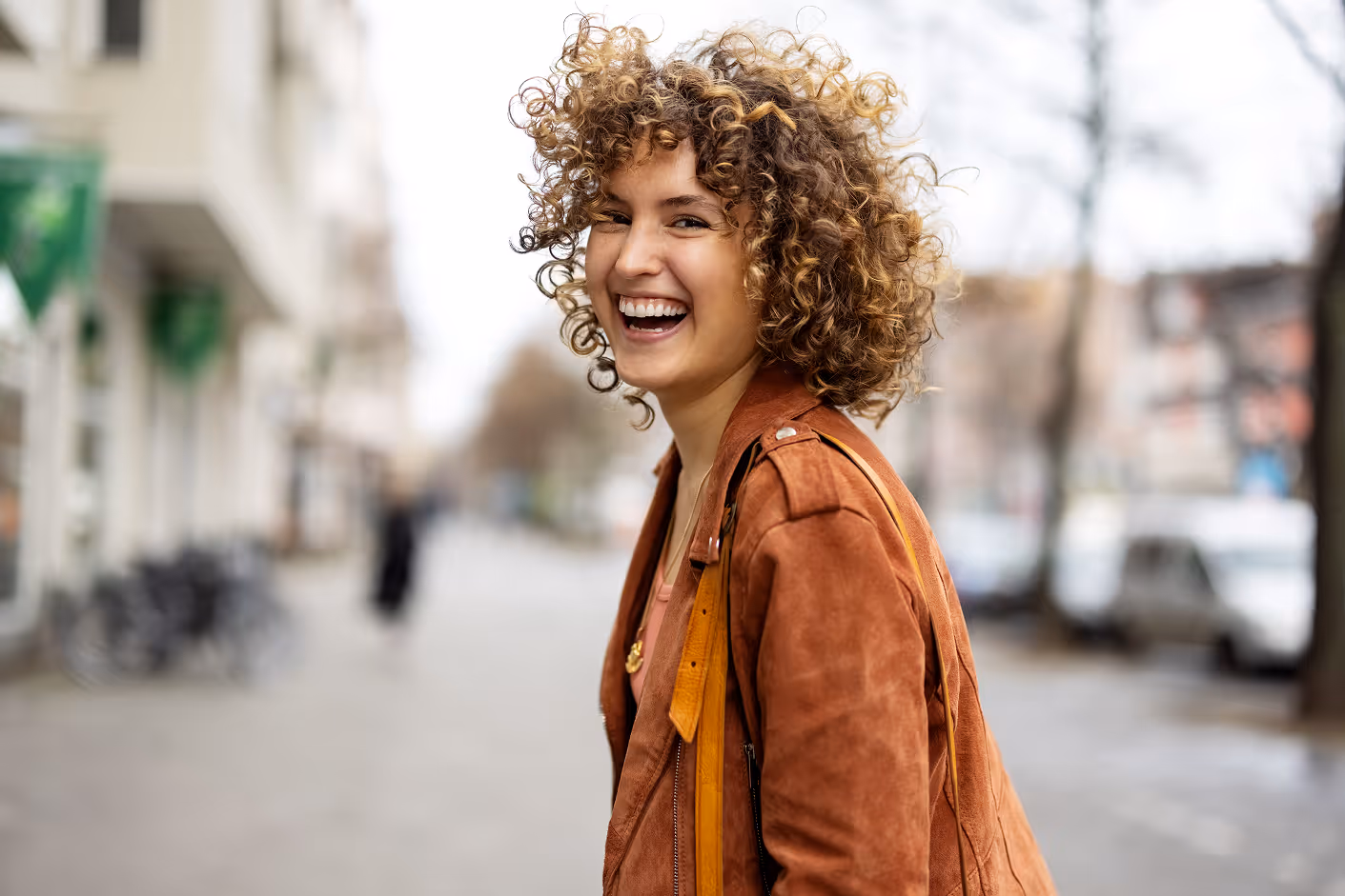 Young woman with curly hair laughing on an urban street wearing a brown jacket.