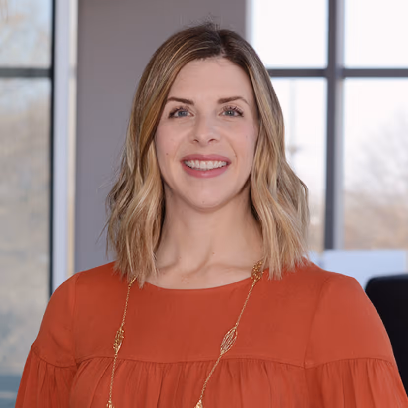 Smiling woman with shoulder-length blonde hair wearing an orange blouse and gold necklace.