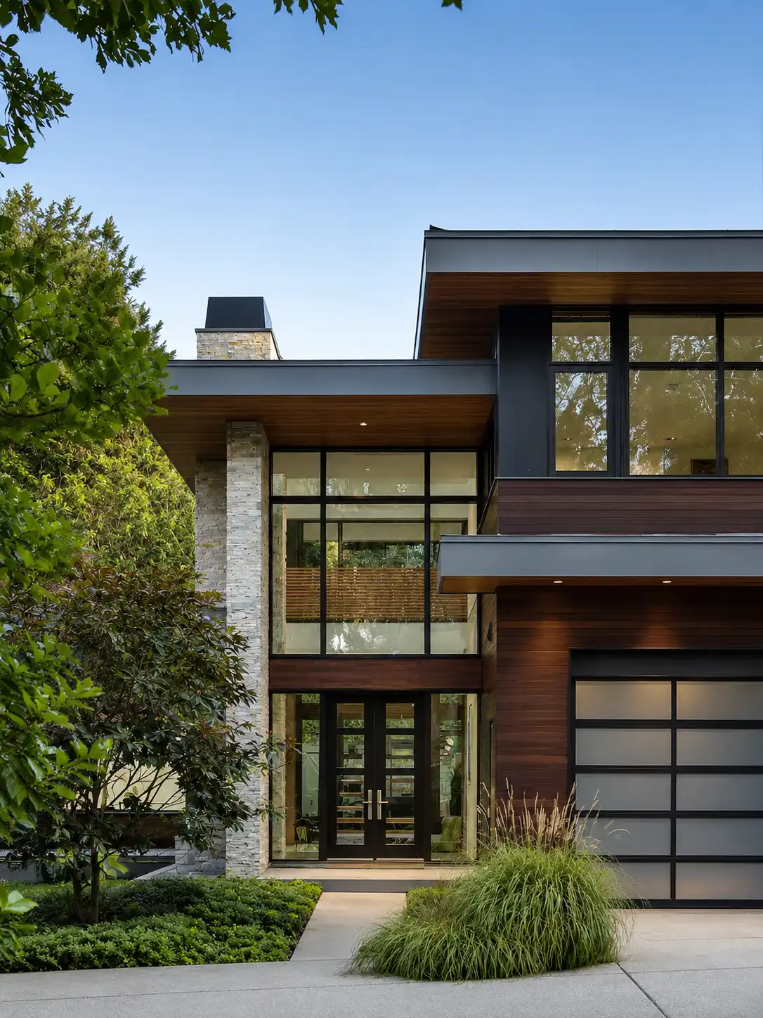 Modern two-story house with large glass windows, wooden accents, stone pillars, and lush greenery.