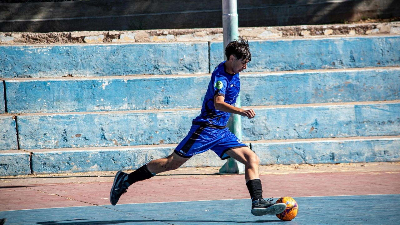 A young soccer player in blue kit mid-action on a futsal court, skillfully controlling an orange ball. The empty blue bleachers form the backdrop.