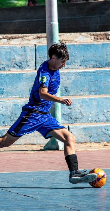 A young soccer player in blue kit mid-action on a futsal court, skillfully controlling an orange ball. The empty blue bleachers form the backdrop.