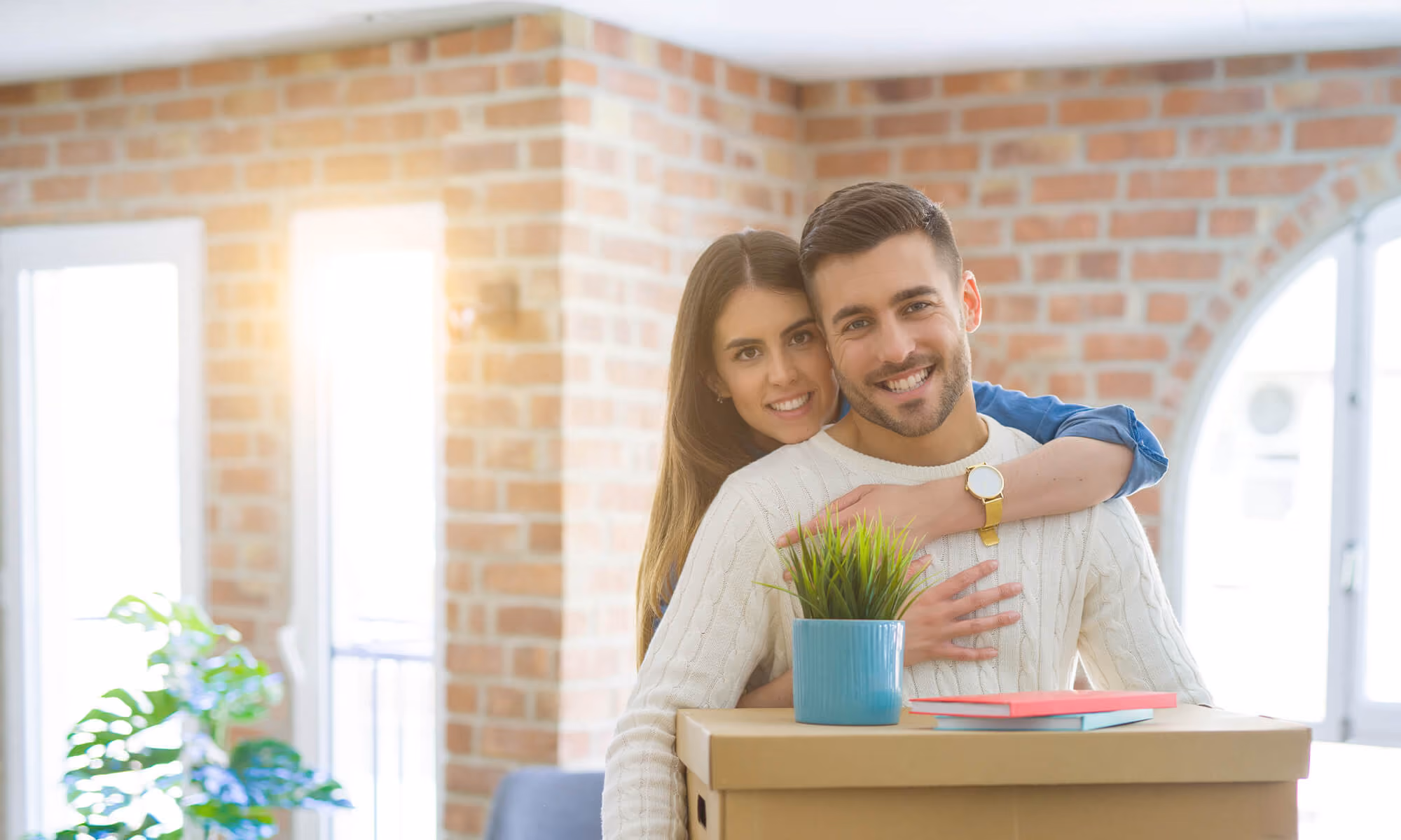 Smiling couple indoors holding cardboard box with potted plant and books, hugging in bright room.