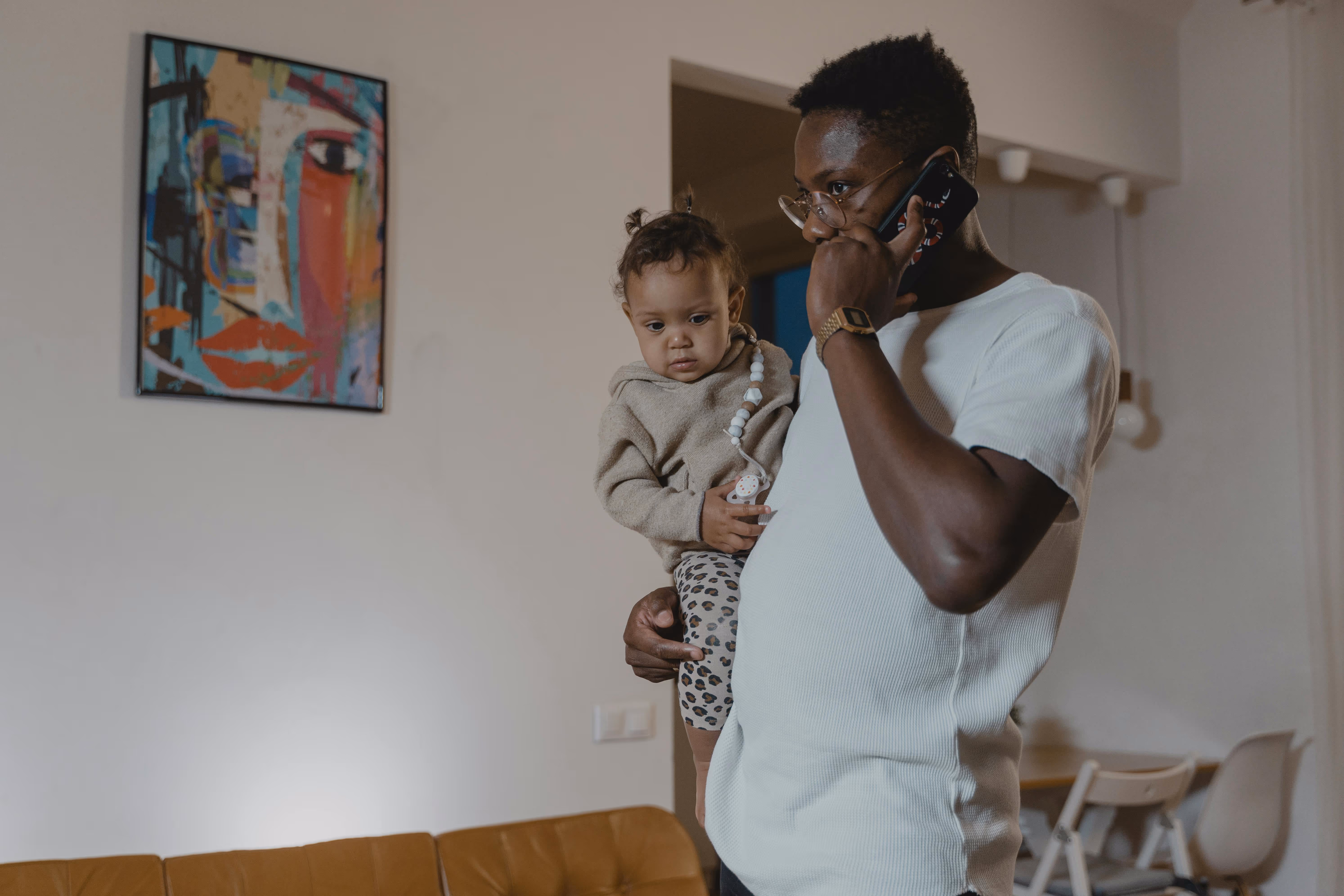 A father holding an infant at home while speaking on the phone with a worried expression