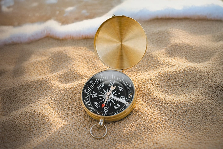A brass compass open on sandy beach showing directions with waves in the background.