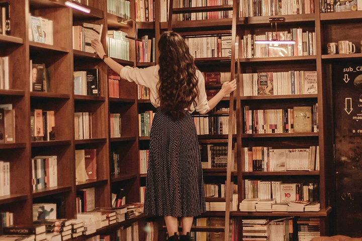 Woman with long hair reaching for a book on a high shelf in a cozy wooden bookstore.