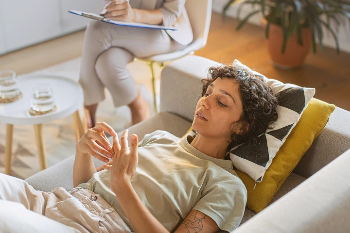 Woman lying on couch talking to therapist taking notes in a bright room with plants.