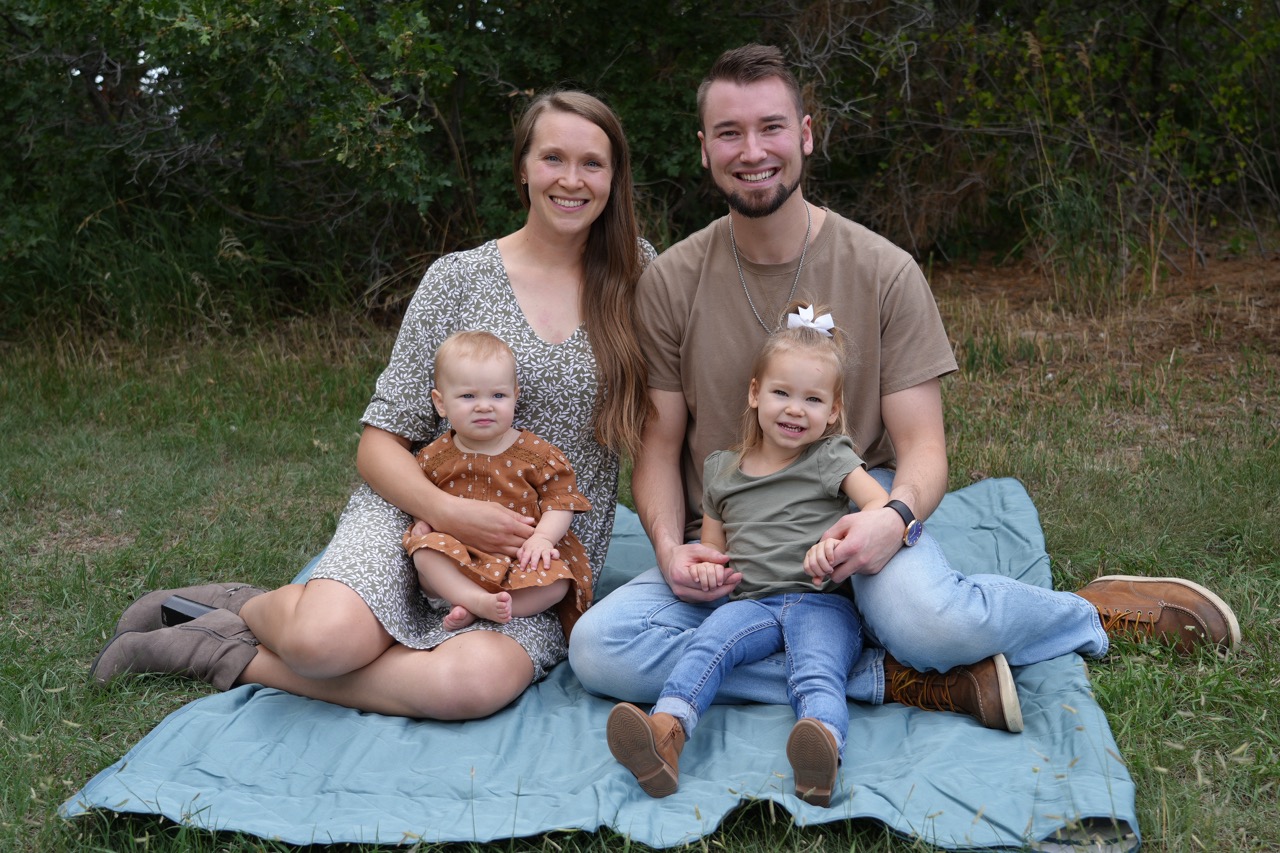 A smiling family of four sitting on a blue blanket outdoors with trees in the background.