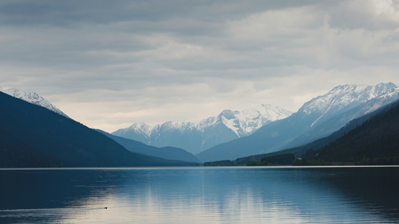 Calm lake reflecting cloudy sky with snow-capped mountains and forested hills in the background.