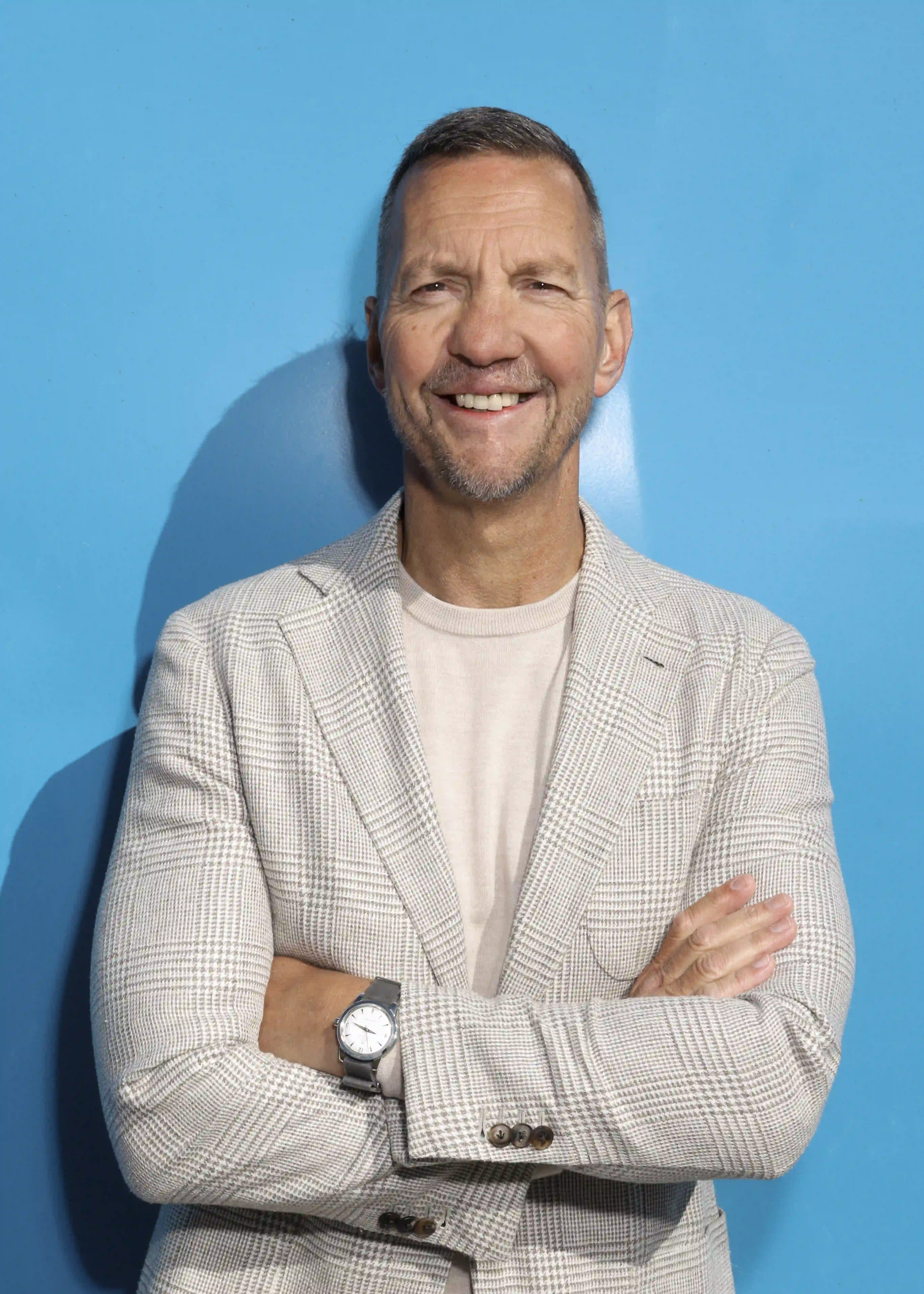 Smiling man with folded arms wearing a beige checked blazer and watch against a blue background. Eddy Beer.