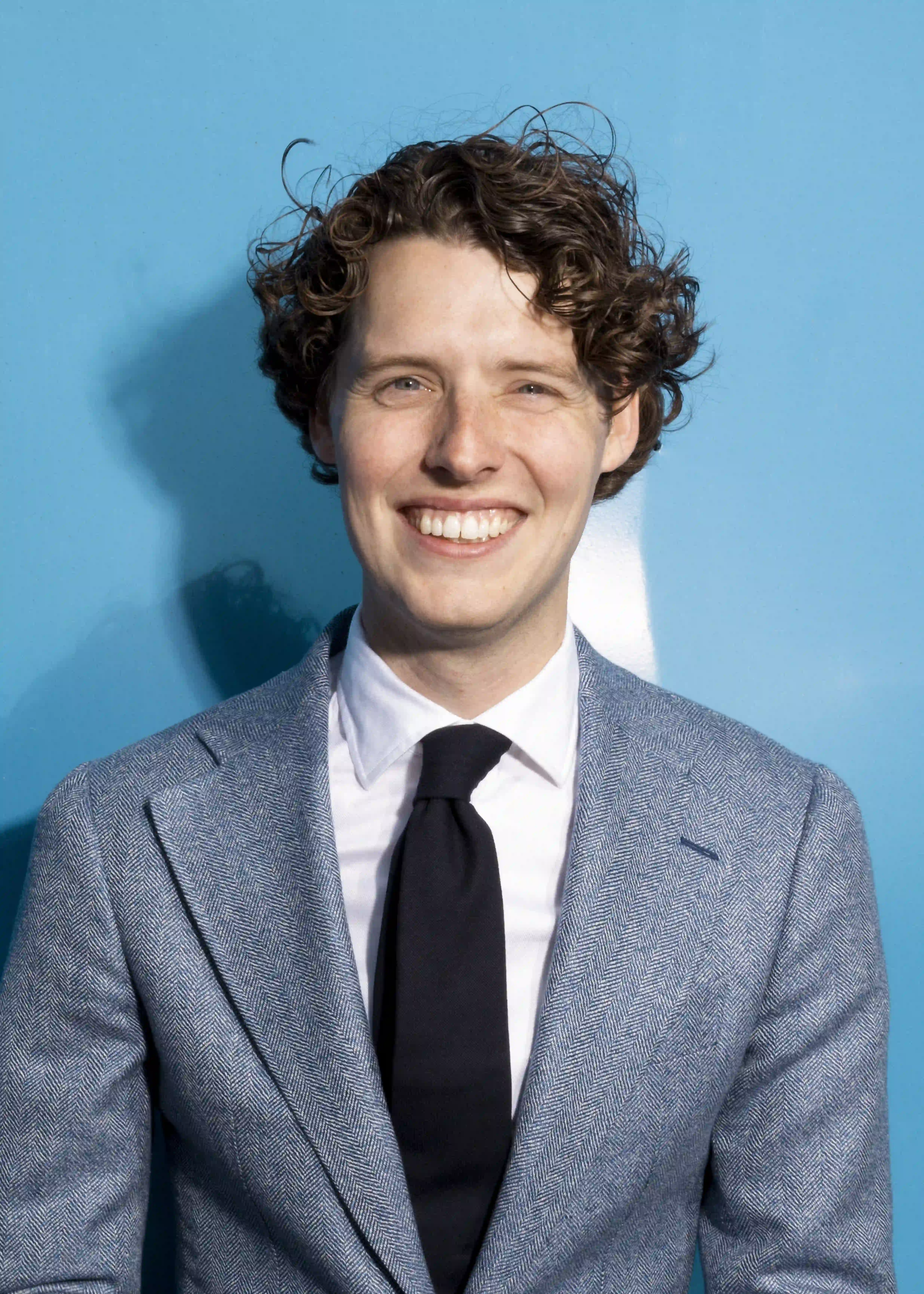 Smiling man with curly hair wearing a blue blazer, white shirt, and black tie against a blue background. Melle Heil.