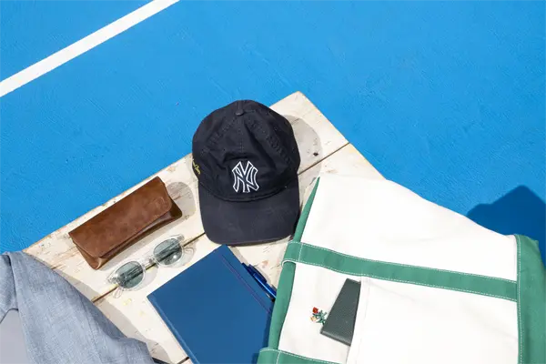 A black cap, sunglasses, brown pouch, and blue notebook on a wooden board by a pool with bright blue water.