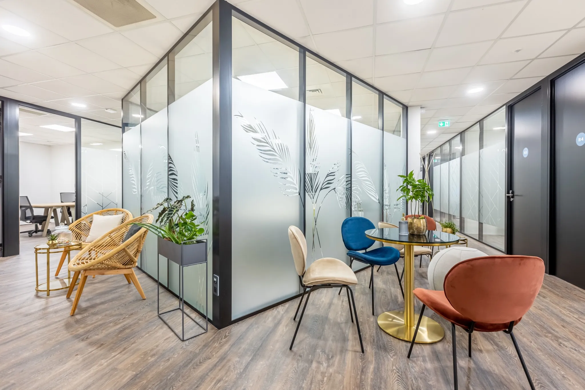 Coin salon avec chaises colorées, table ronde dorée, fauteuils en rotin, et plantes vertes.
