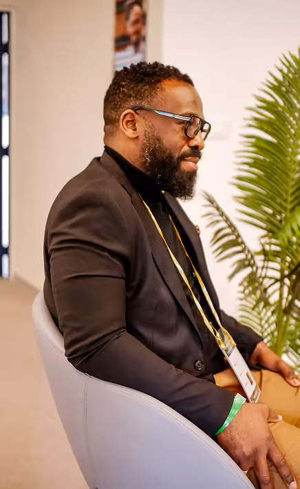 Homme portant des lunettes et un blazer noir assis sur une chaise grise près d'une plante verte.