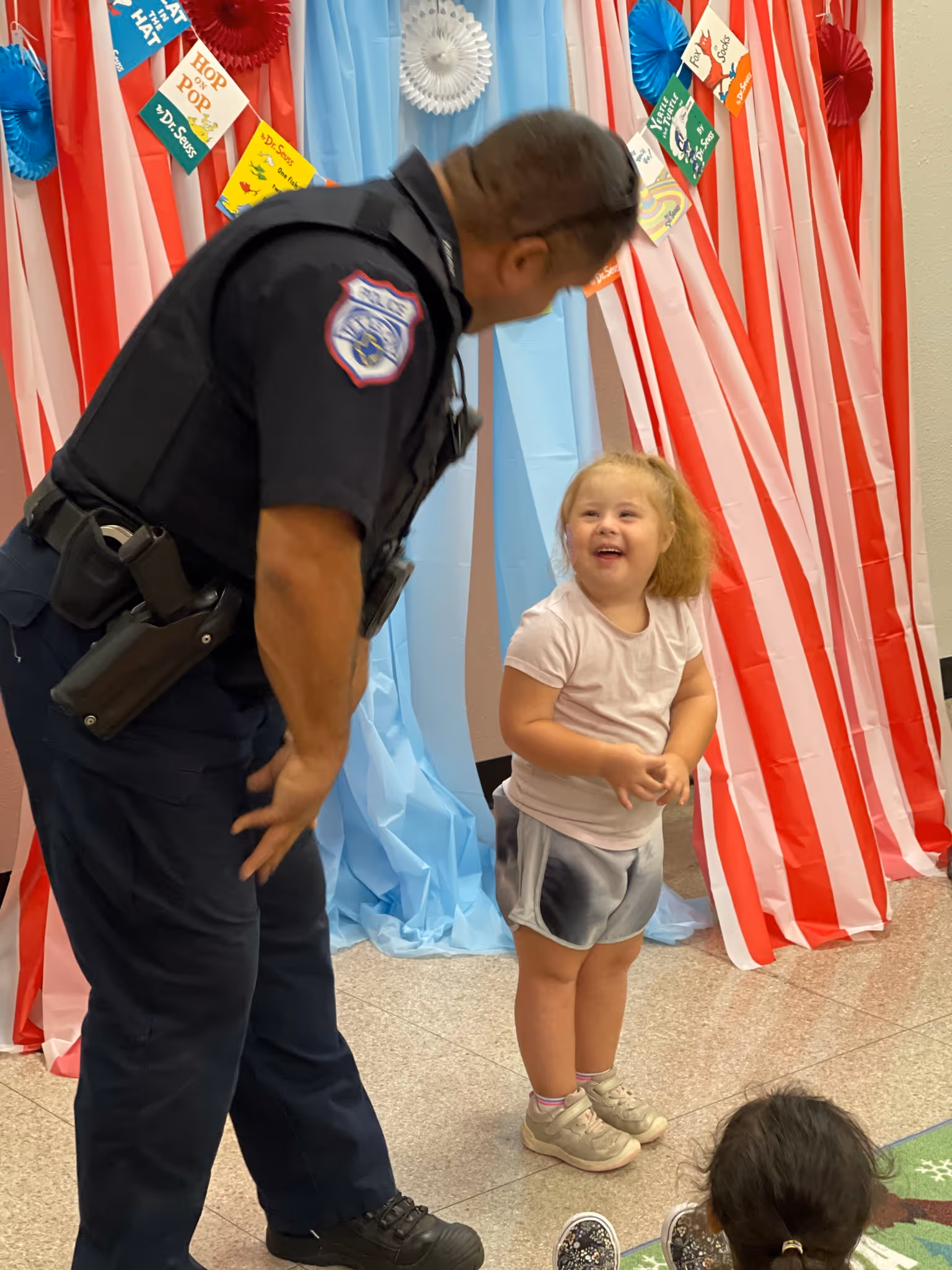 Police officer bending down and smiling at a smiling young girl in a decorated room.