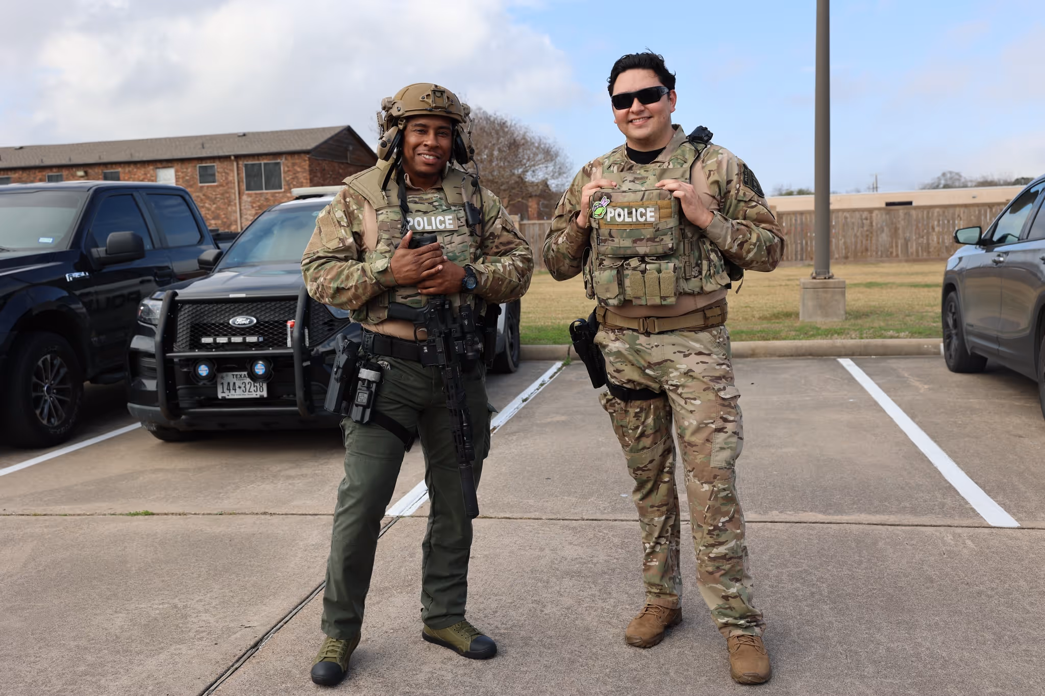 Two armed police officers in tactical gear standing in a parking lot with police vehicles behind them.
