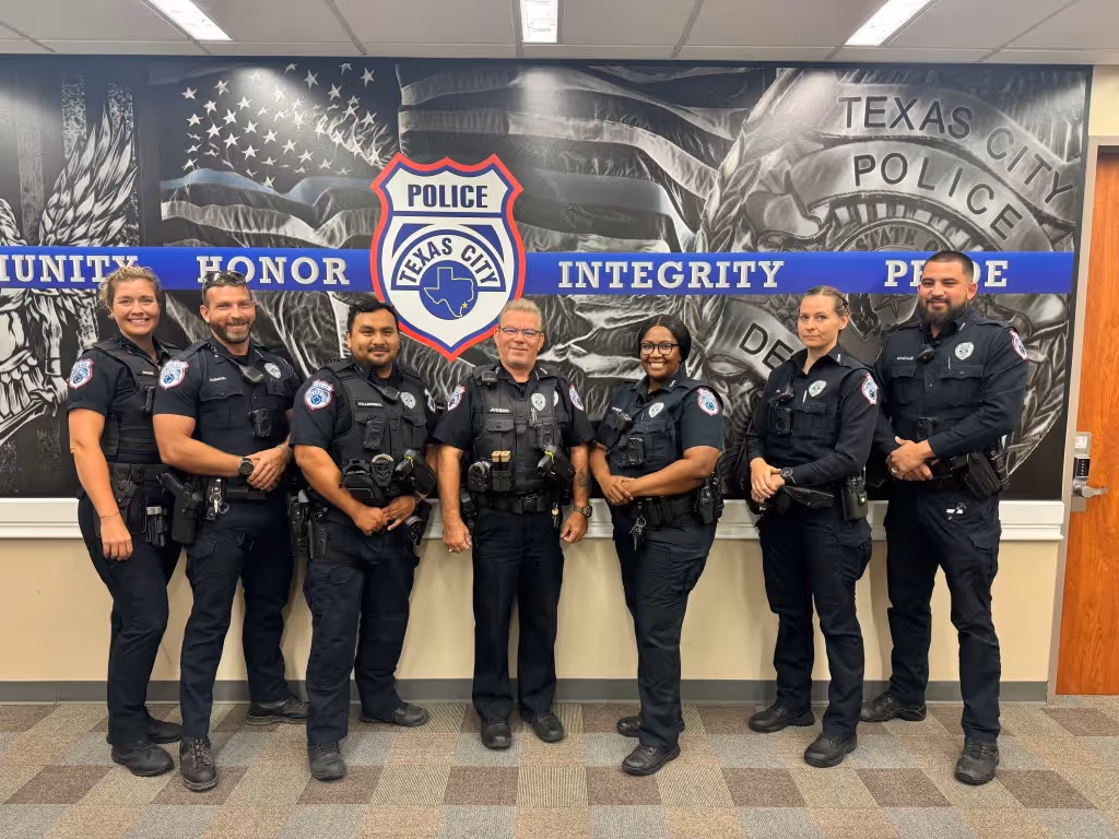 Seven Texas City police officers standing in front of a wall with police badge mural and words honor, integrity.