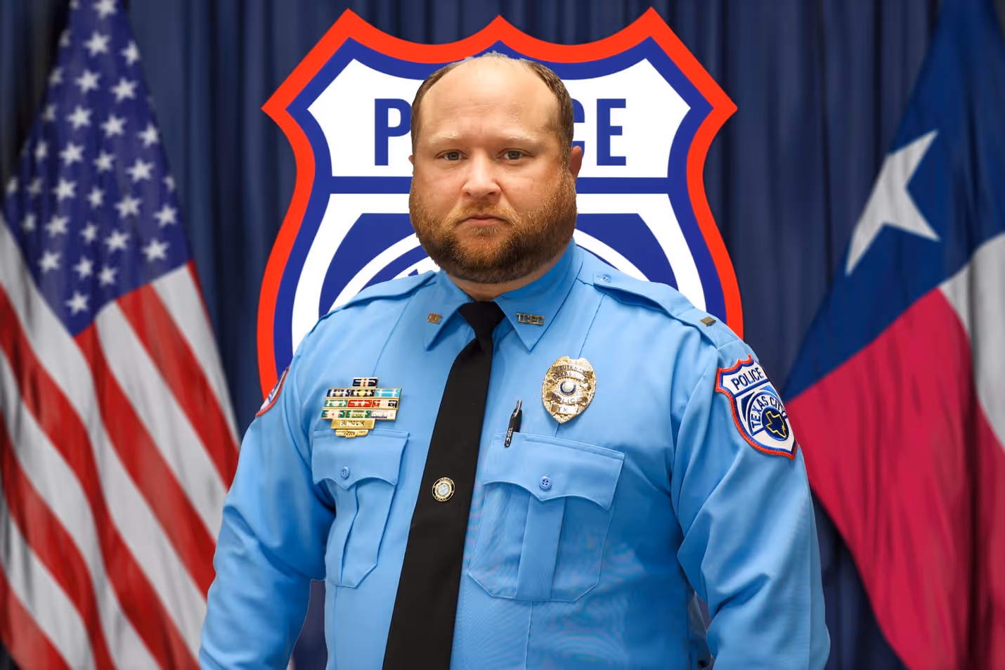 Bearded male police officer in blue uniform stands with American and Texas flags behind him.