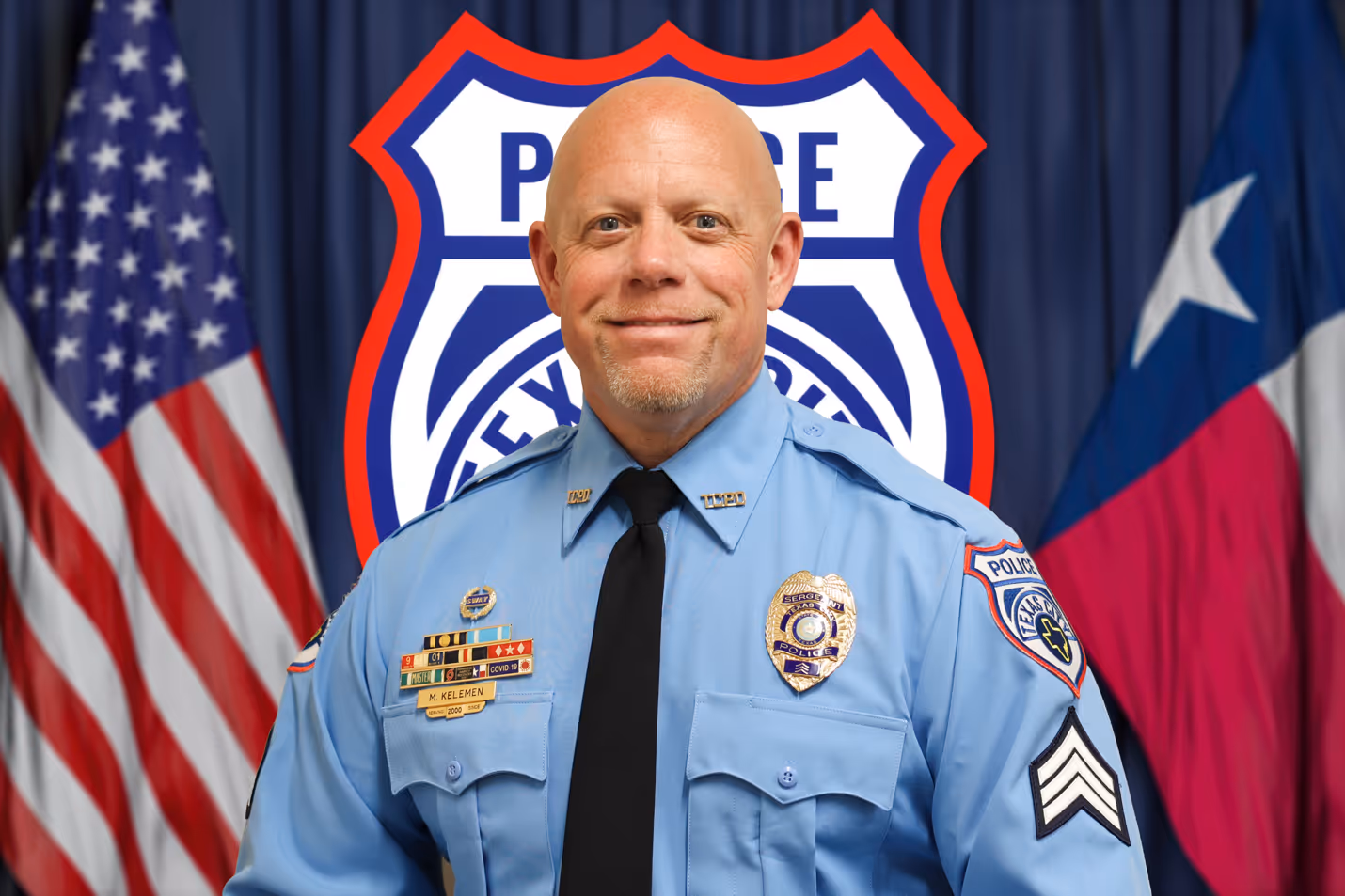 Smiling bald police sergeant in blue uniform with badges, standing before American and Texas flags.