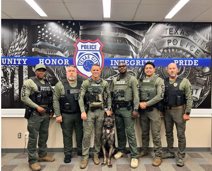 Six Texas City police officers in uniform with police dog in front of a mural with police badge and values.