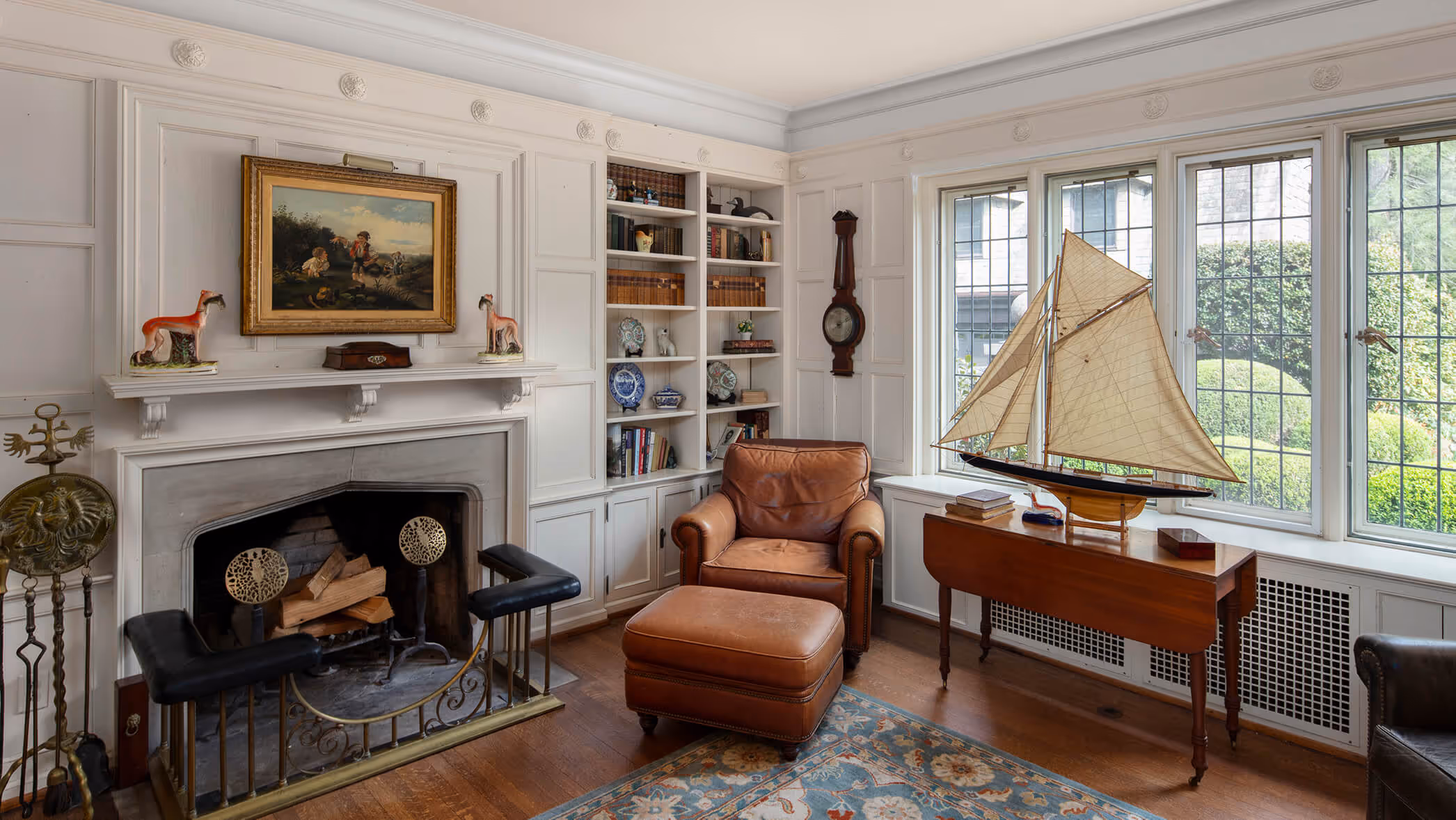 Cozy living room with leather armchair, fireplace, bookshelf, model sailboat on wooden table, and large window.