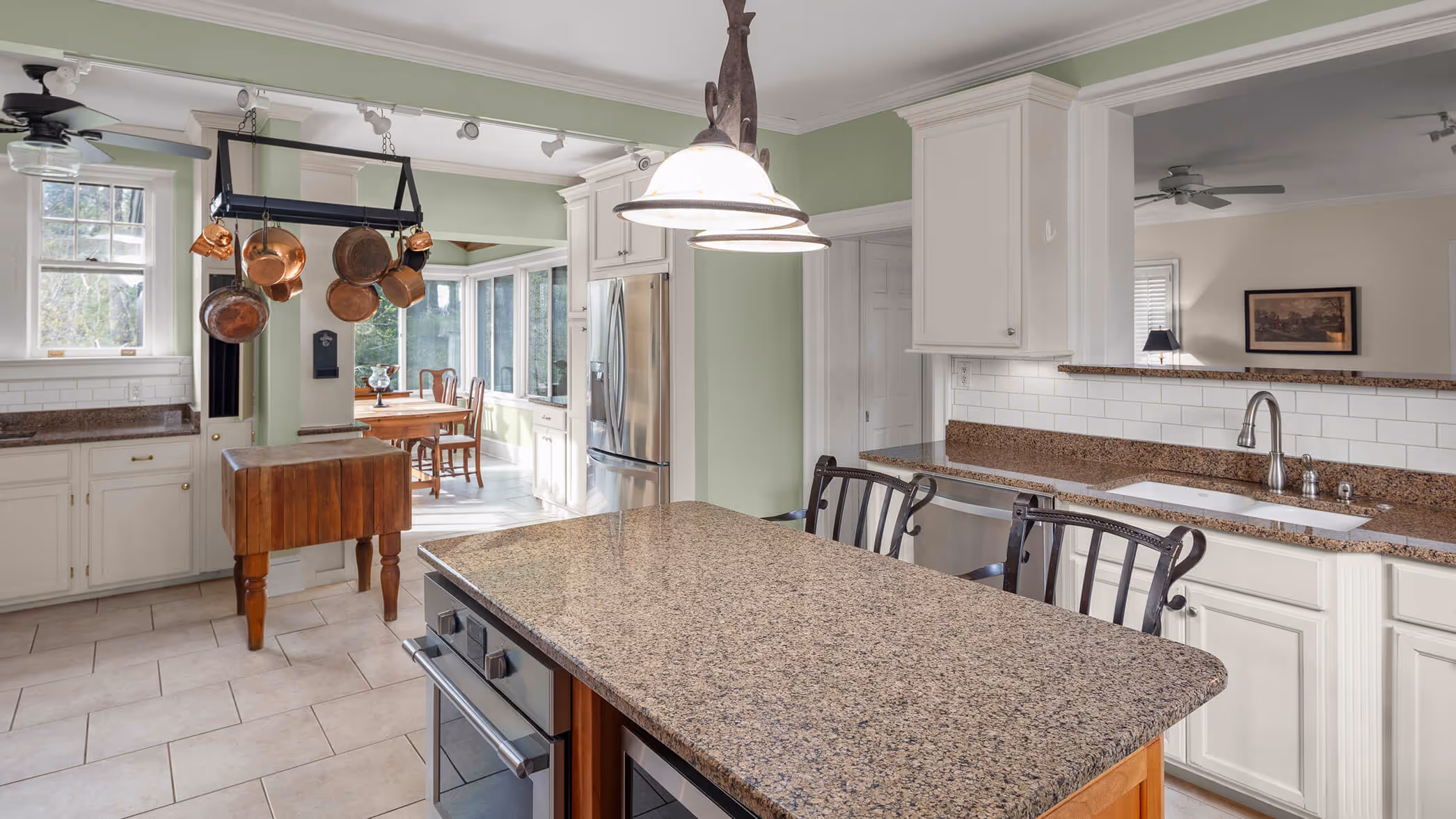 Spacious kitchen with granite island, hanging copper pots, white cabinets, and dining area with large windows.