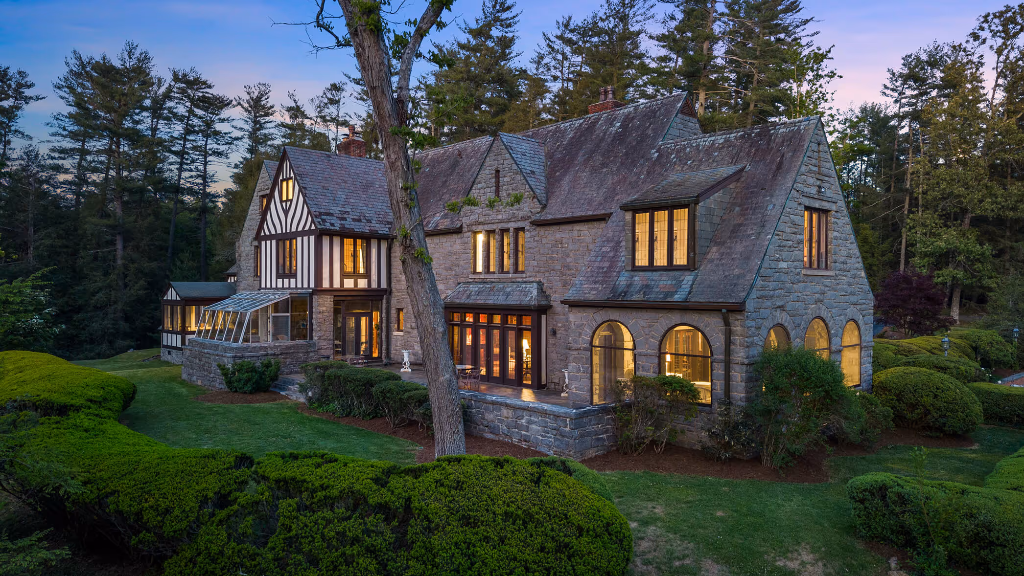 Stone Tudor-style house with lit windows surrounded by green bushes and trees at dusk.