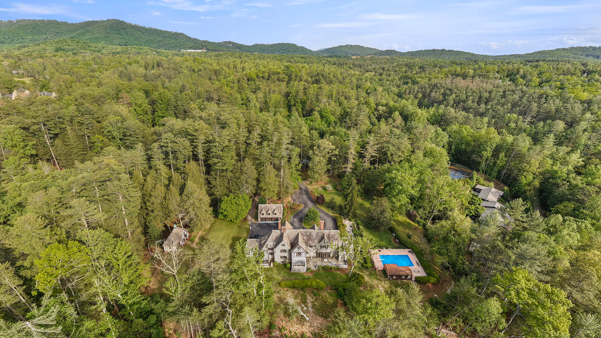 Large estate with stone house, pool, and tennis court surrounded by dense forest and hills.