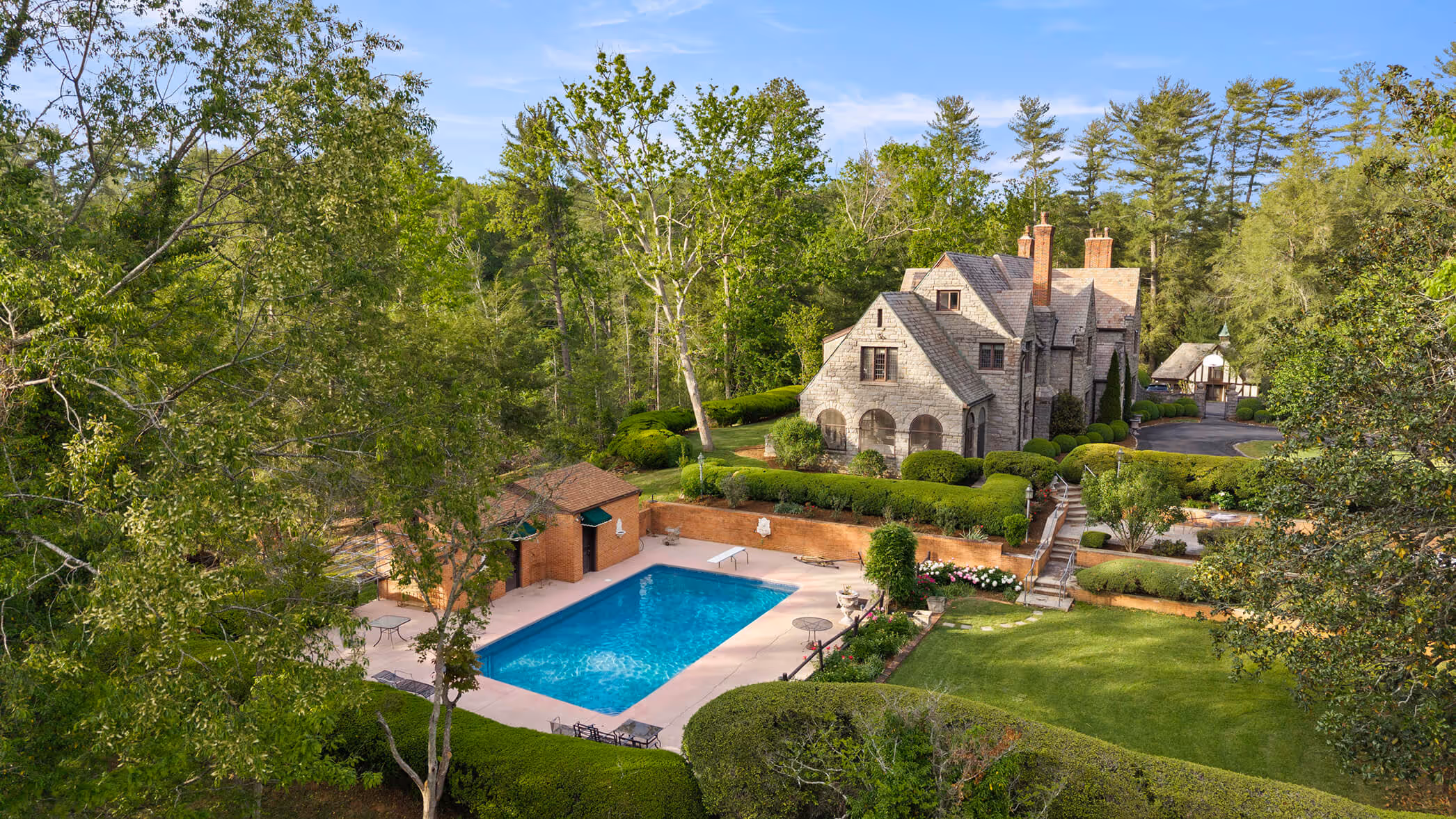 Large stone house with trimmed bushes near a rectangular pool surrounded by trees and lawn.