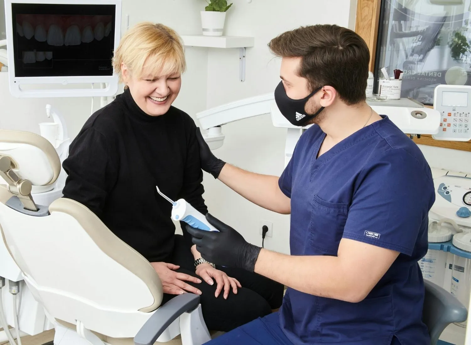 Dentist in blue scrubs and black mask talking to a smiling female patient sitting in a dental chair.