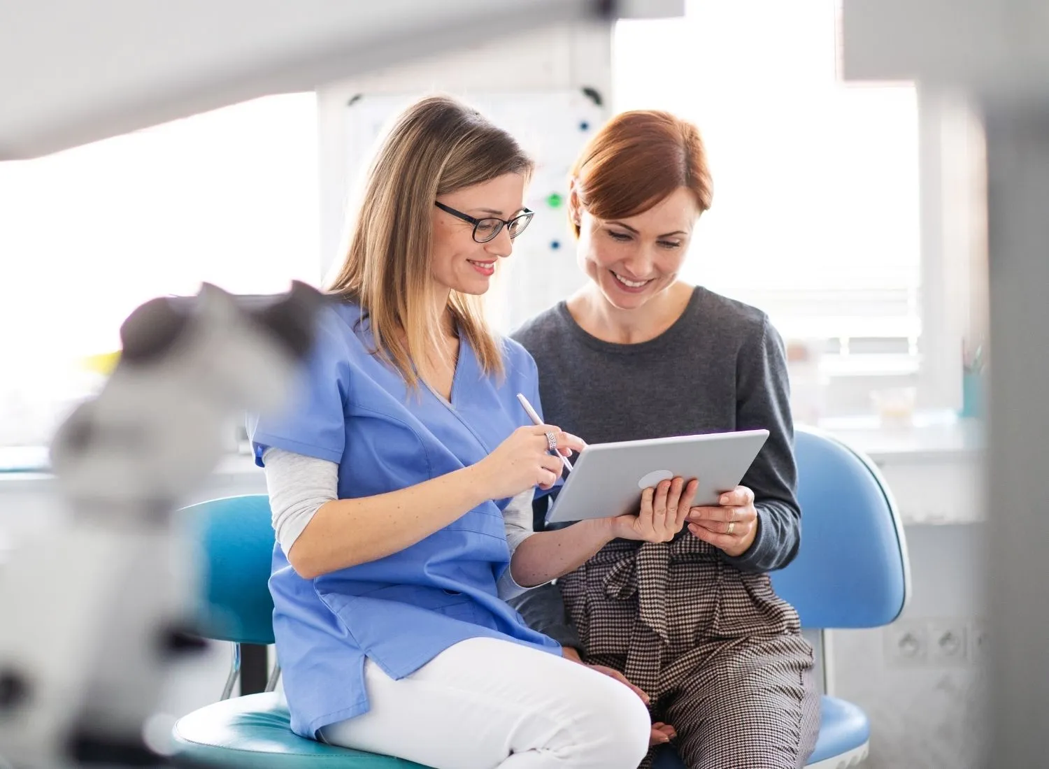 Smiling female healthcare professional in blue scrubs showing tablet to smiling woman in gray sweater.