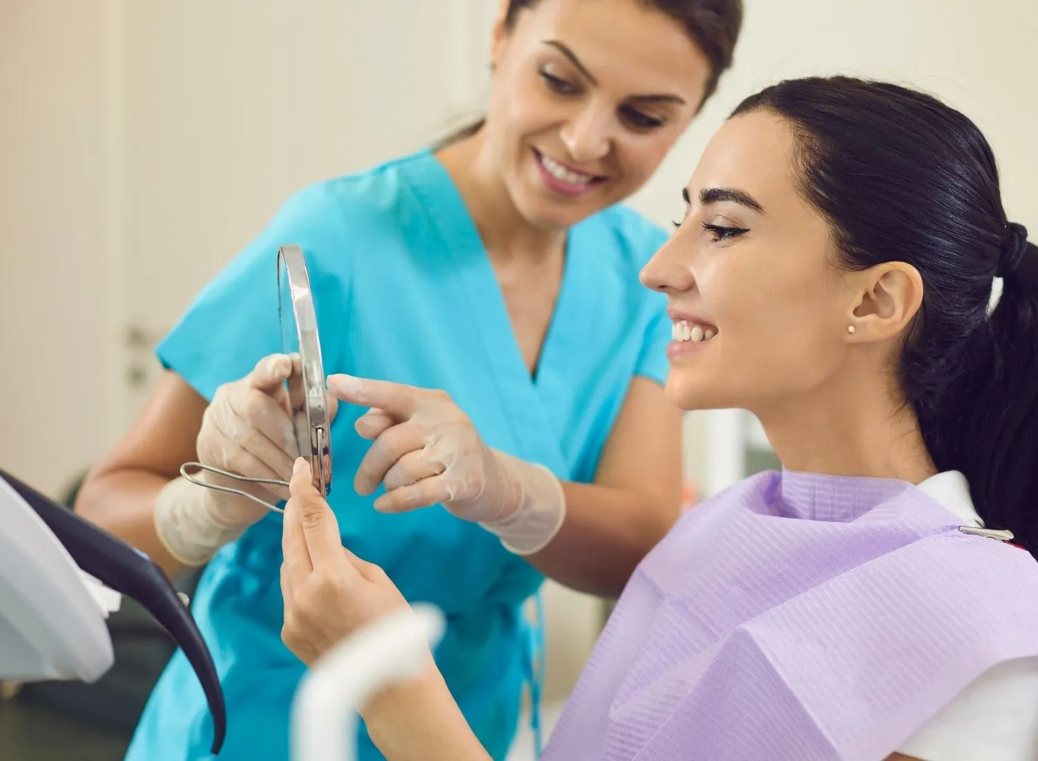 Dentist explaining dental care to a smiling female patient holding a handheld mirror.