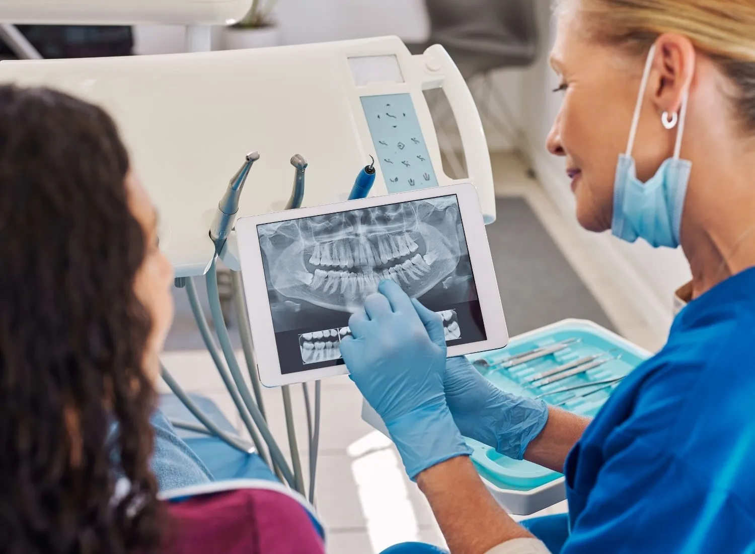 Dentist wearing gloves and mask showing dental X-ray on tablet to patient in clinic.