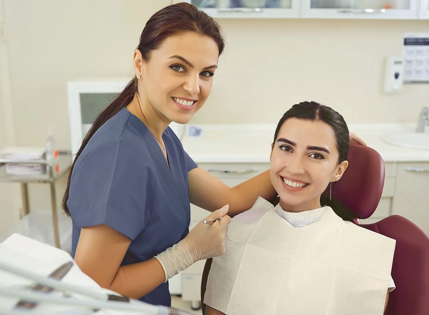 Smiling female dentist in scrubs and gloves with a happy patient in a dental chair.