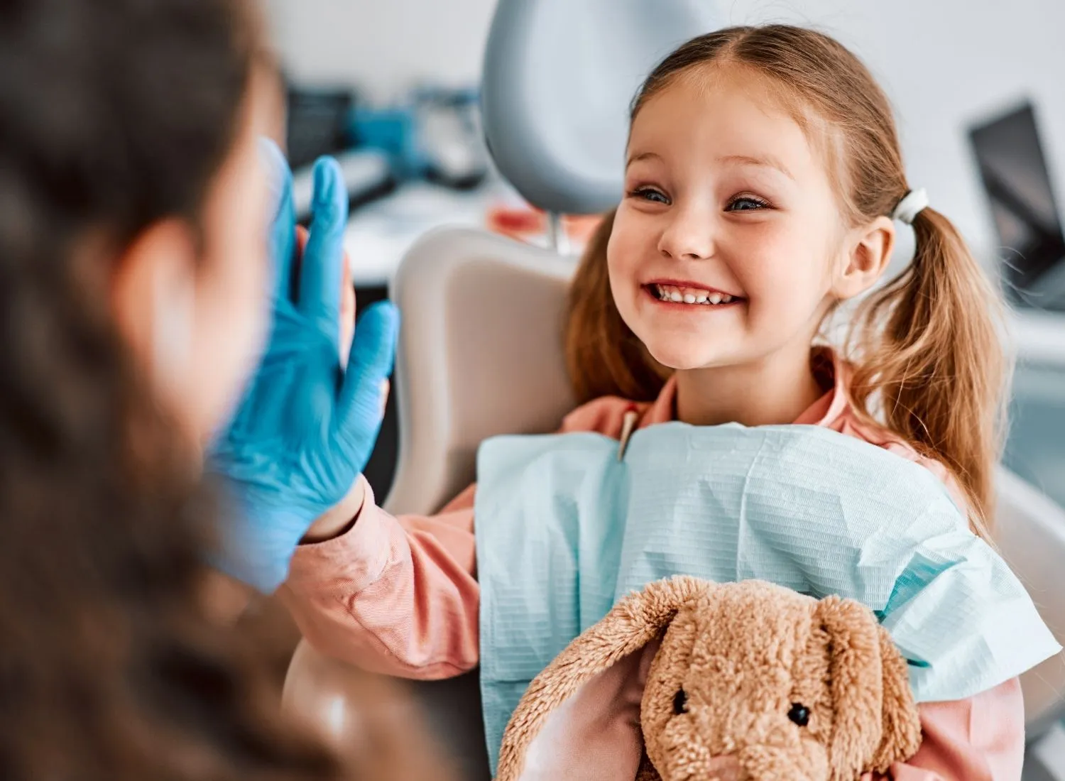Smiling little girl in dental chair with bib holding a plush bunny and high-fiving a gloved hand.