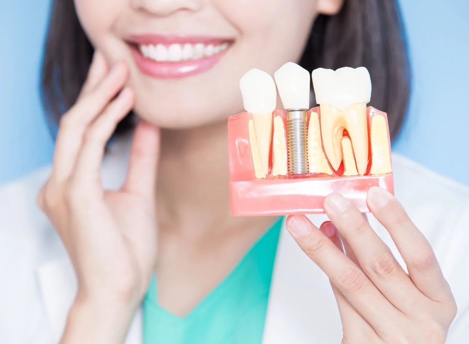 Dentist holding dental model showing natural teeth and a dental implant, smiling with hand on jaw.