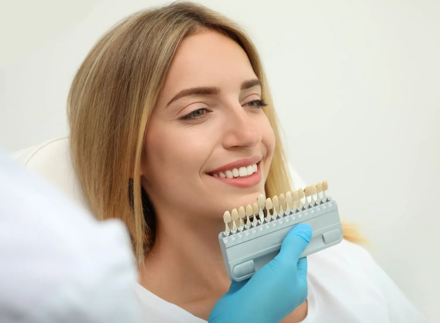 Dentist wearing blue gloves holds a tooth shade guide next to a smiling woman's teeth.