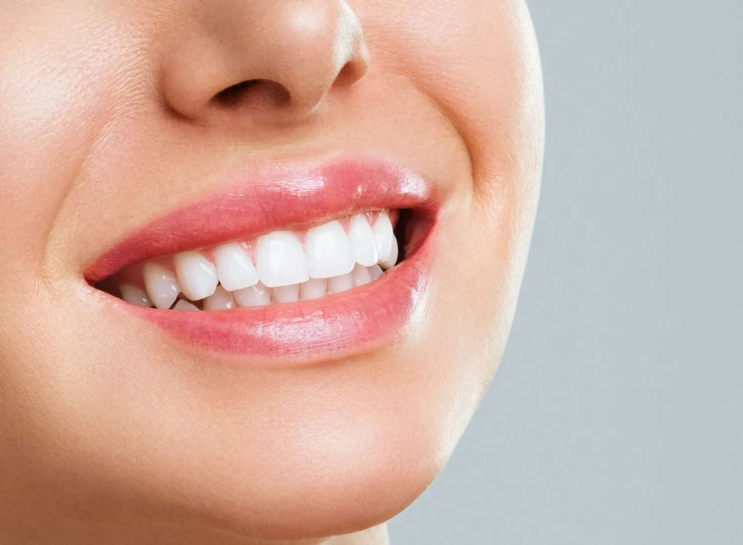 Close-up of a smiling mouth showing clean, white teeth and pink lips on a light gray background.