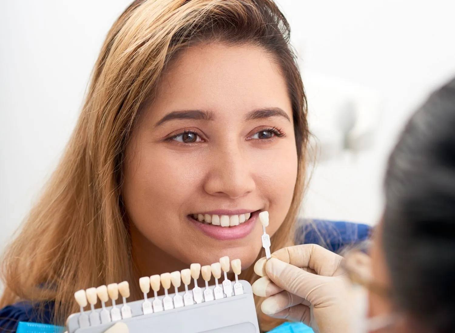 Dentist comparing dental shade guide to smiling woman’s teeth for color matching.