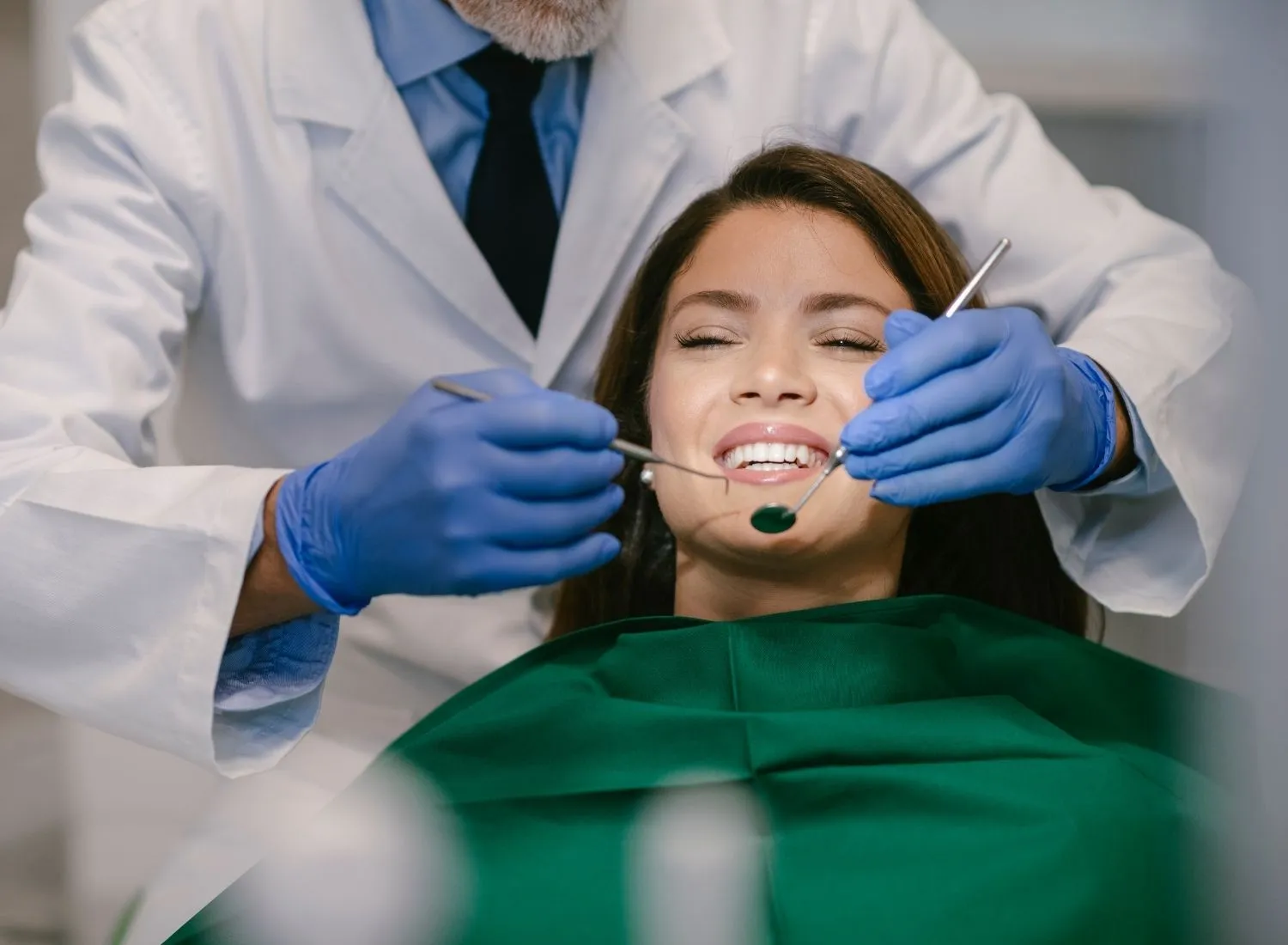 Dentist wearing gloves examining a smiling woman's teeth with dental tools in a clinic.