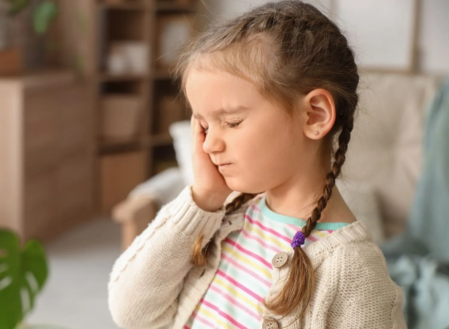 Young girl with braided hair holding her cheek with eyes closed, showing pain or discomfort.