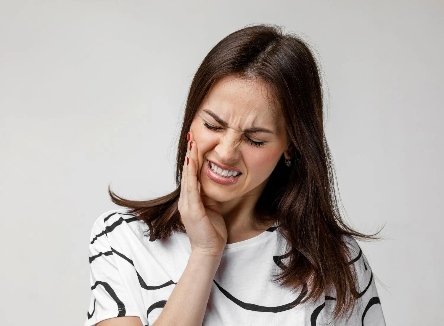 Woman with dark hair wearing white shirt holds her cheek with a painful expression.