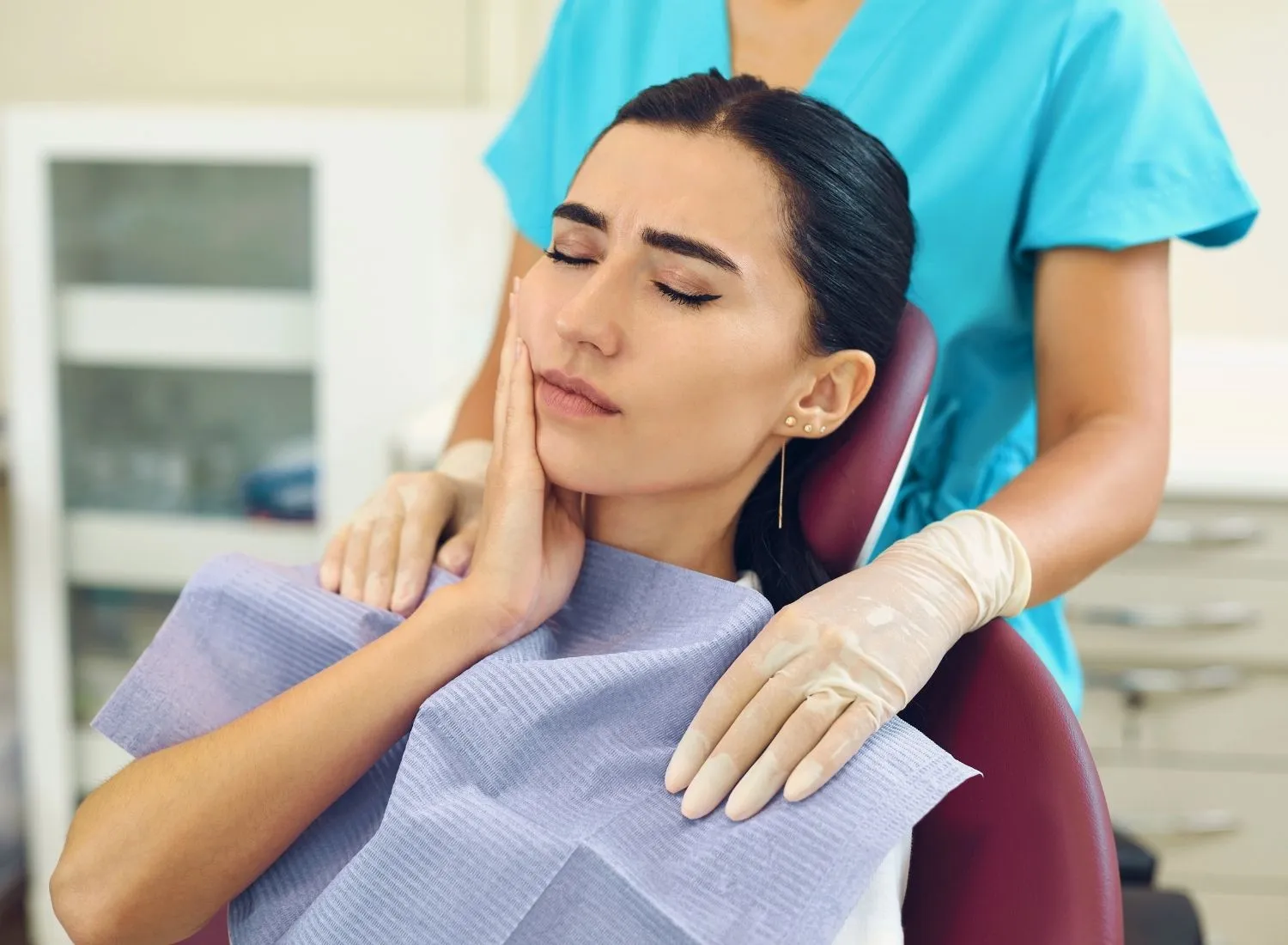 Woman in dental chair holding her cheek in pain while a gloved dental professional comforts her.