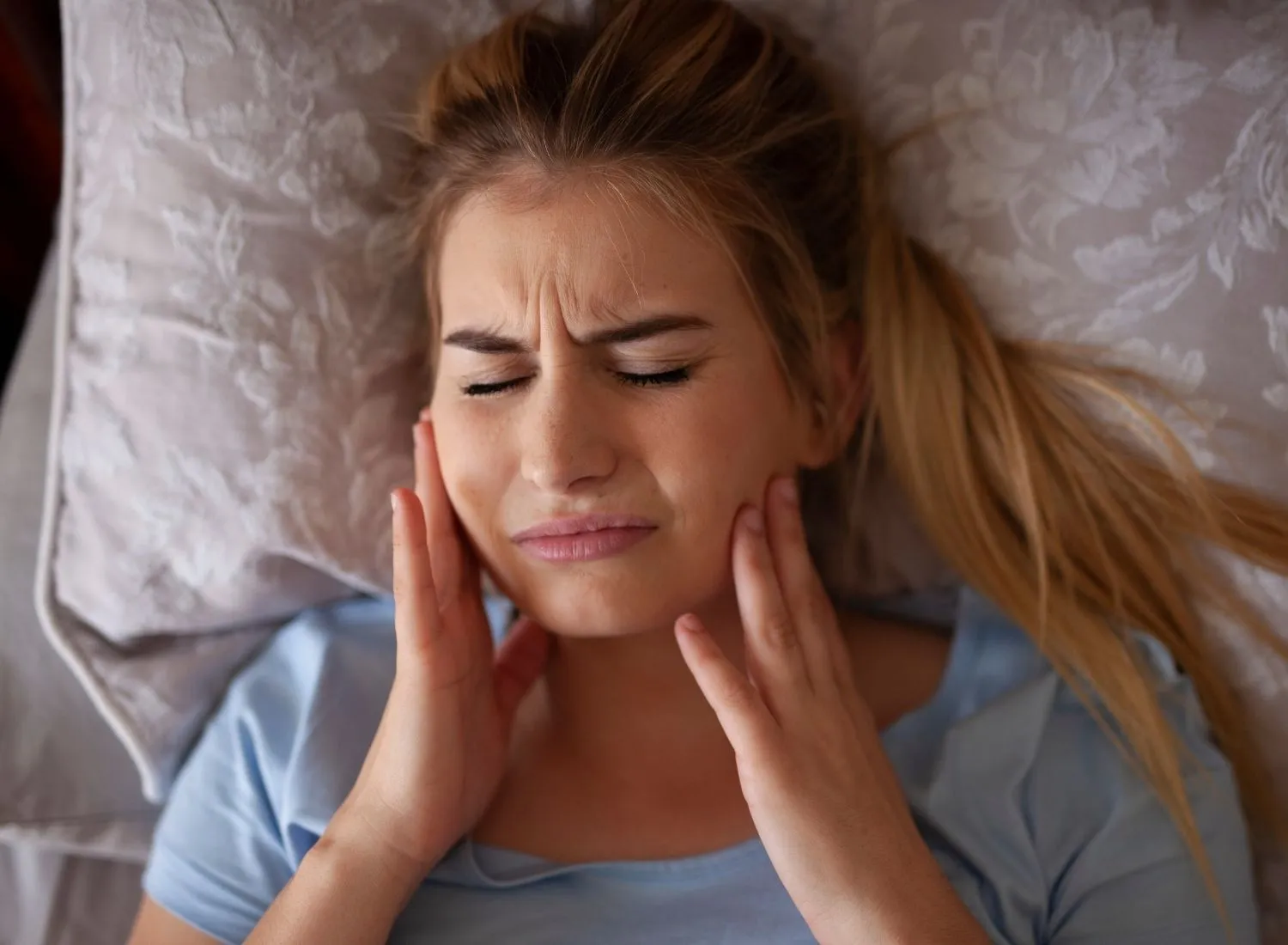 Young woman lying in bed with eyes closed, holding her jaw in pain from tooth or jaw discomfort.