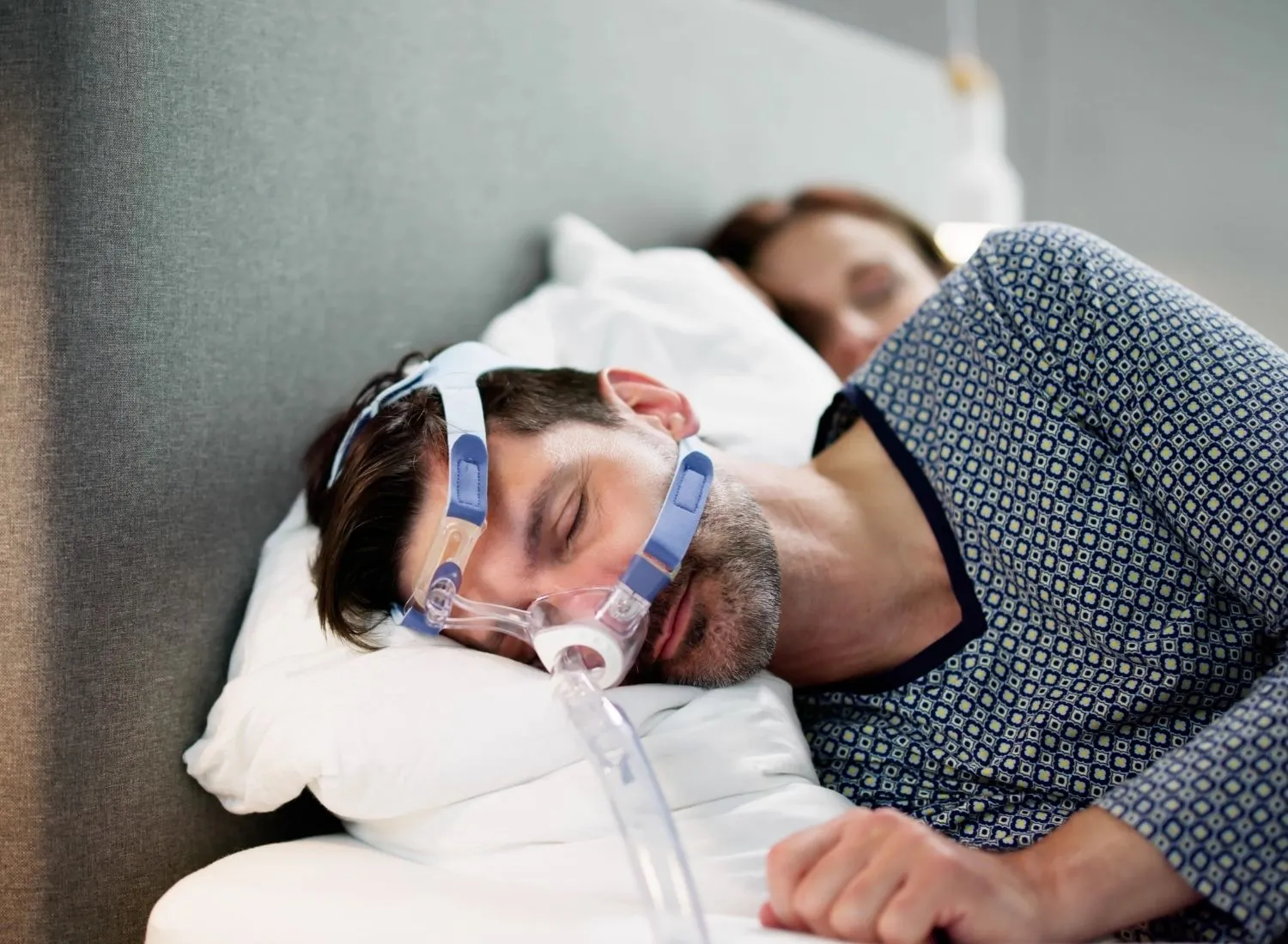 Man sleeping with a CPAP mask on in bed next to a woman also asleep.