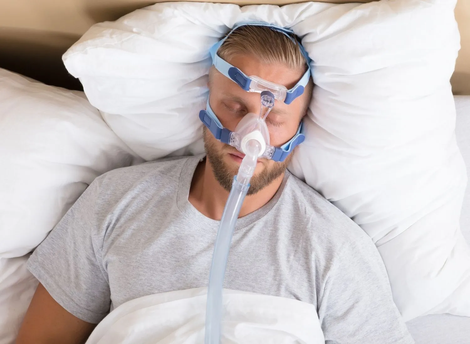 Man sleeping with CPAP mask on his face, lying on white pillows and covered with a white blanket.