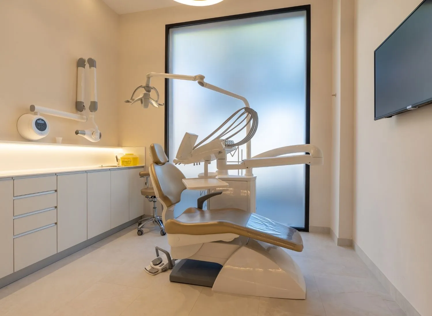 Modern dental clinic room with a beige dental chair, equipment, and a large frosted window.