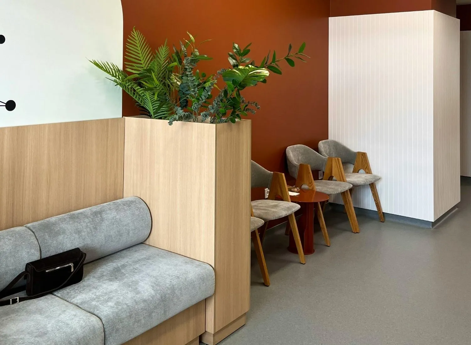 Modern waiting area with gray cushioned seats, wooden chairs, small table, and a planter with green plants.